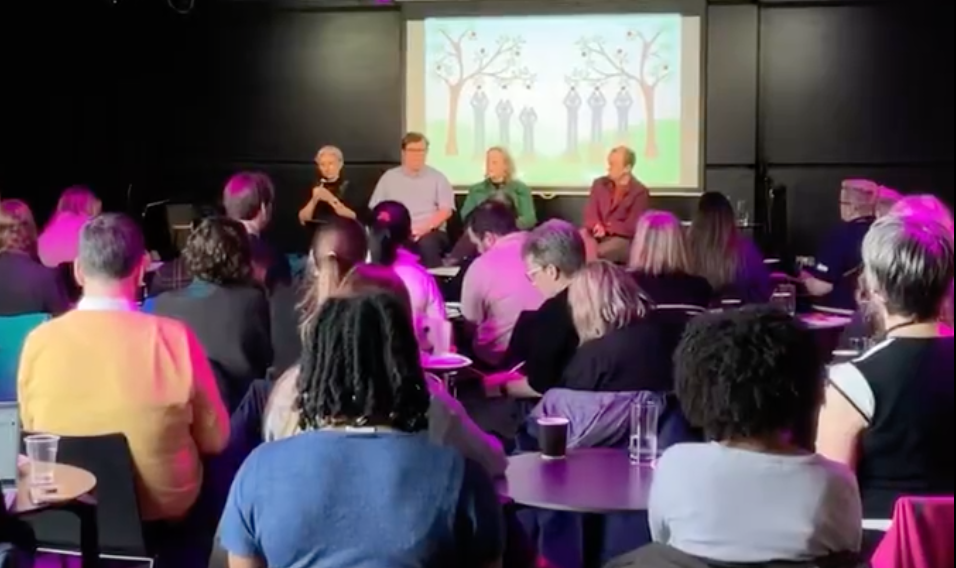 Panel of speakers on a stage with a crowd assembled in front of them and a slide behind them showing the difference between equality and equity.