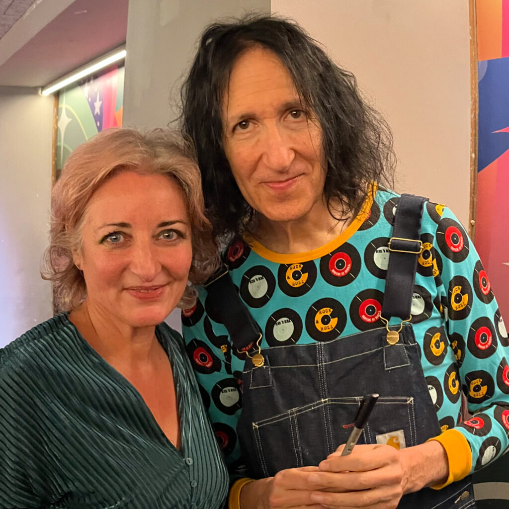 Image of Rebeca and Gordon together at the event. Rebecca is wearing an aquamarine dress; Gordon is wearing dungarees over a turquoise top with large coloured spots
